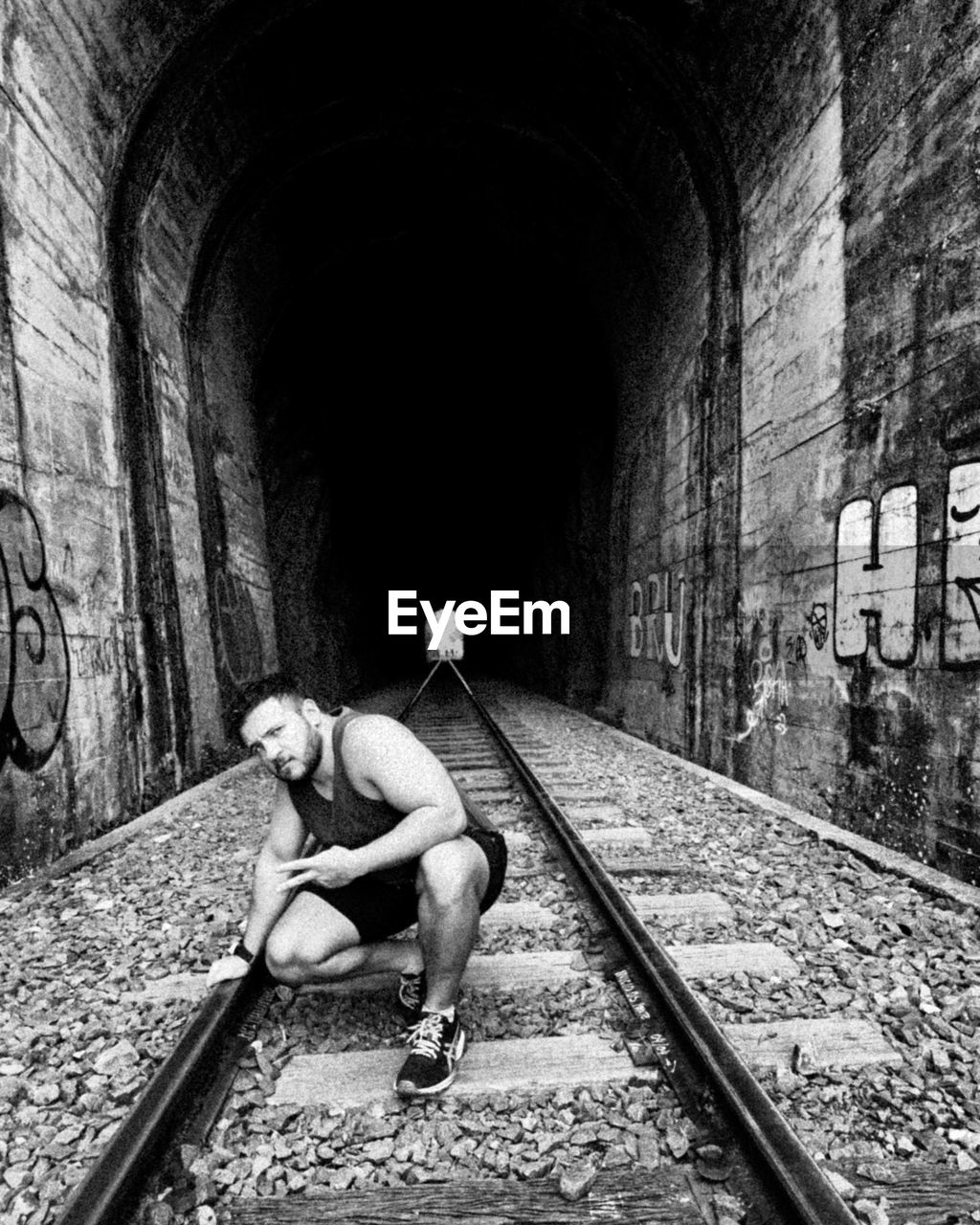one person, track, railroad track, full length, rail transportation, architecture, black and white, adult, tunnel, monochrome, infrastructure, lifestyles, arch, monochrome photography, built structure, transportation, men, person, public transportation, day, sitting, casual clothing, leisure activity, young adult, wall - building feature, graffiti