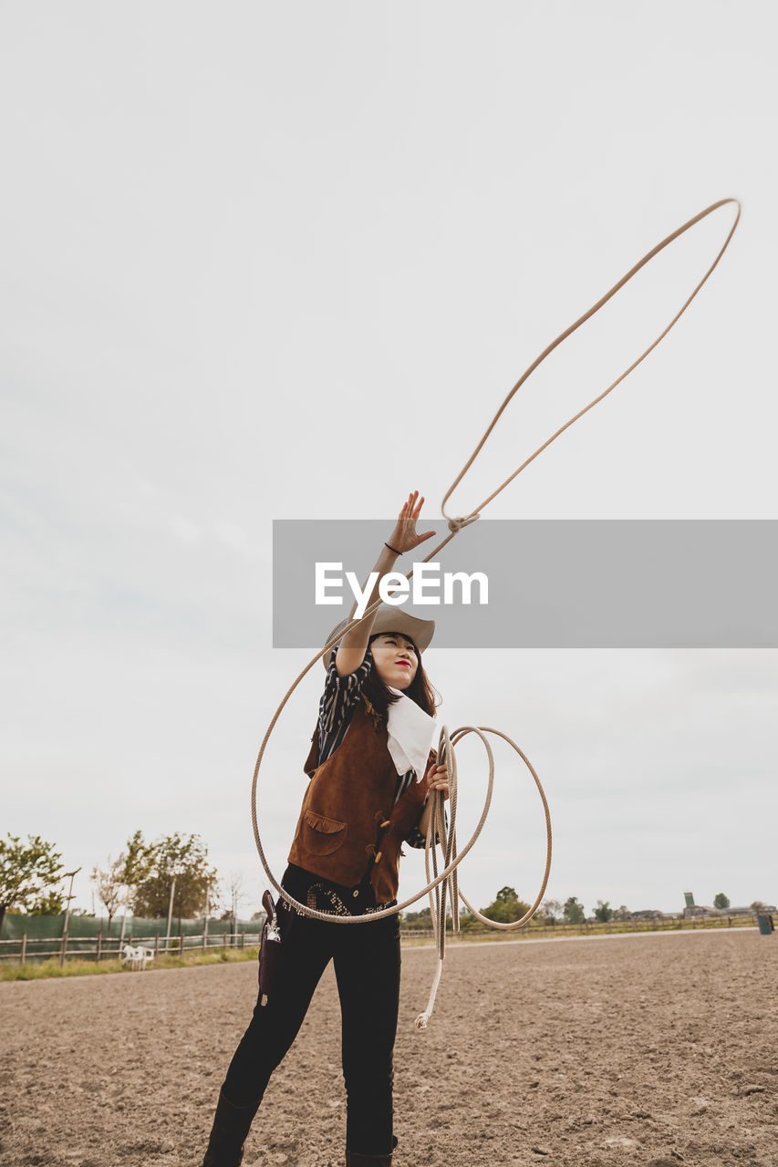 Cowgirl throwing lasso while standing at ranch against sky