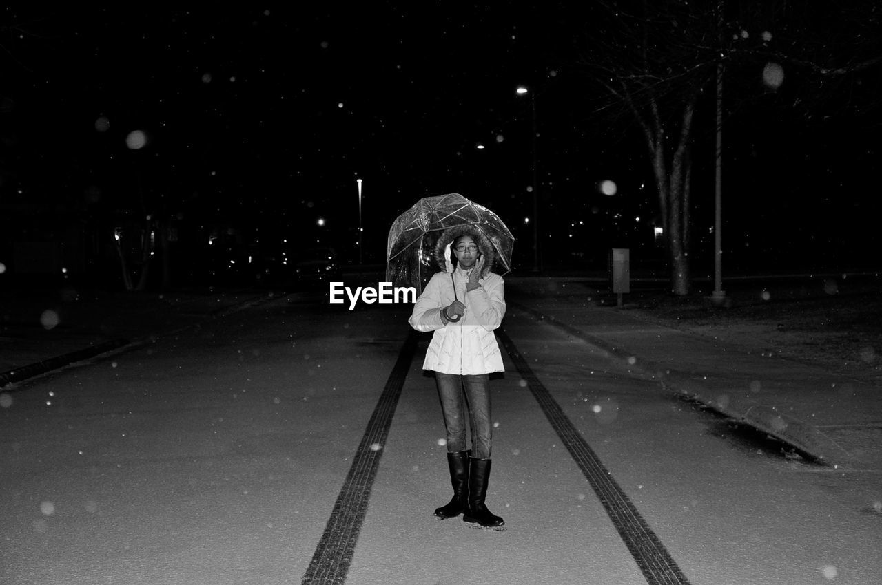 Portrait of young woman holding umbrella while standing on street at night during winter