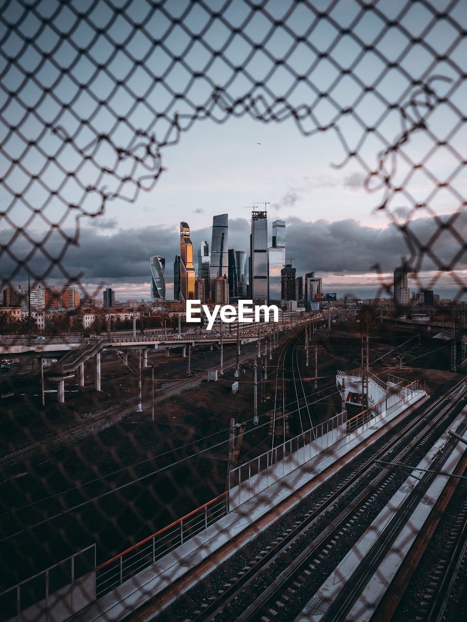 Railroad tracks seen through fence against cityscape during sunset