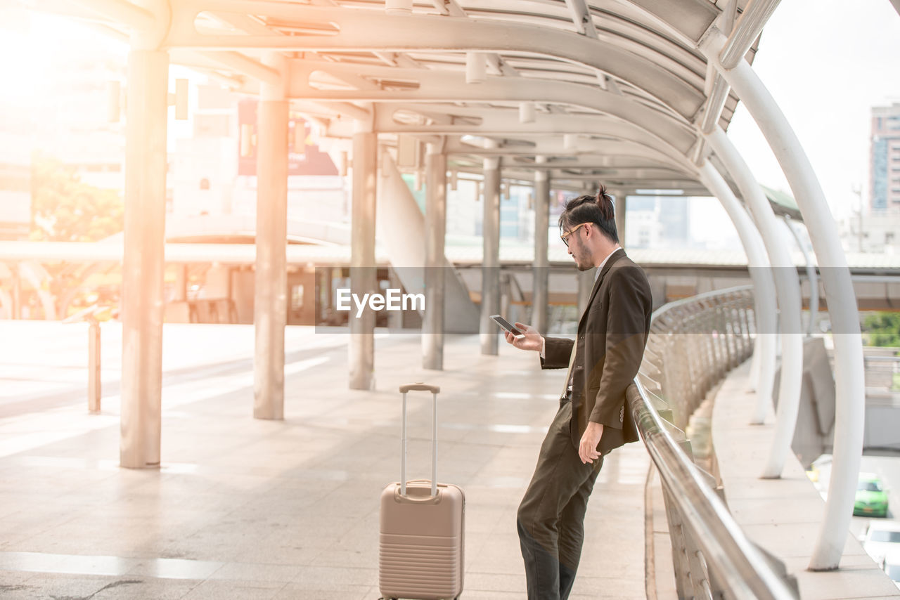 SIDE VIEW OF MAN USING MOBILE PHONE WHILE WALKING ON RAILWAY STATION