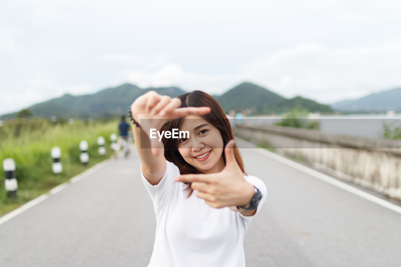 Portrait of smiling young woman making finger frame while standing on road