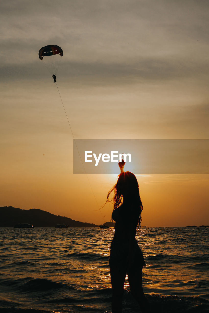 Girl pointing at the paraglide in the sky at the sunset in summer. silhouette of people concept.