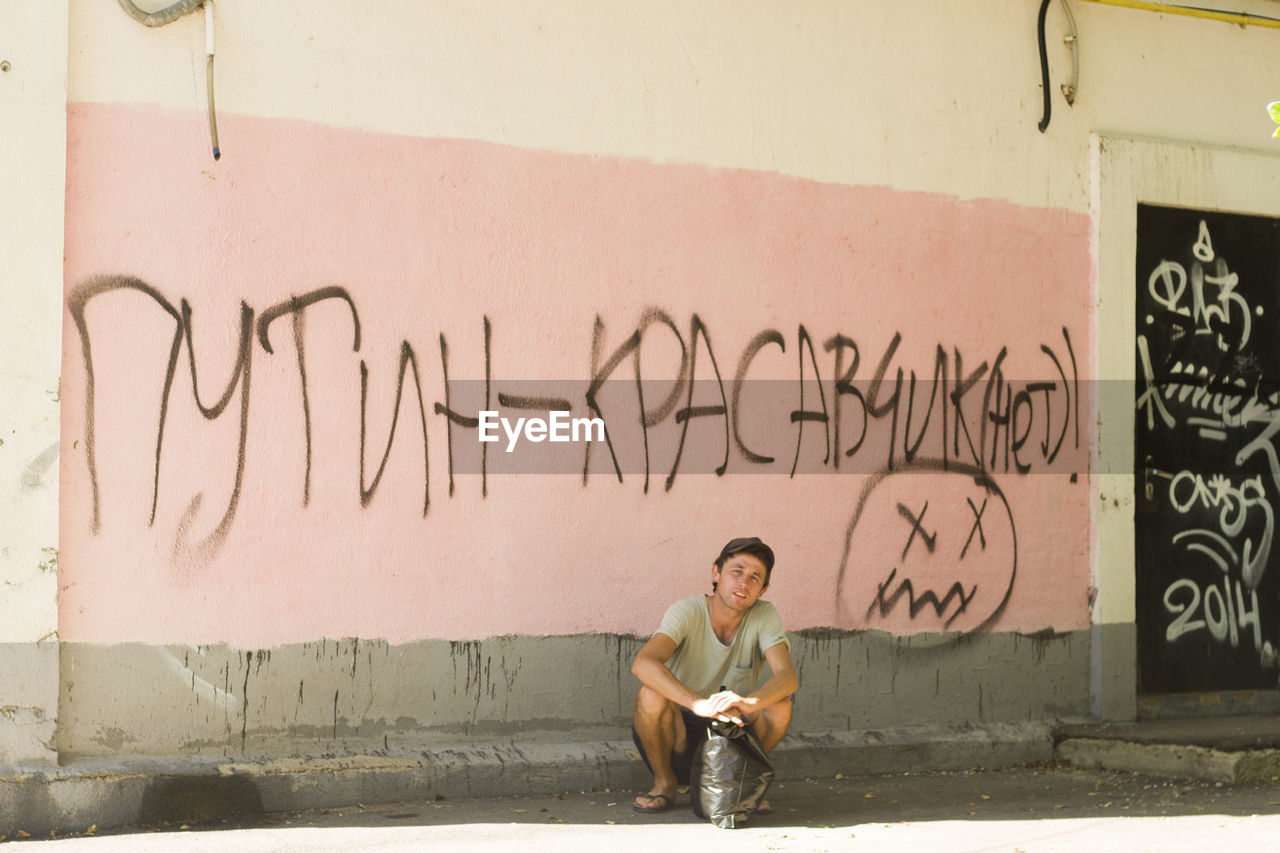 Full length of man crouching against graffiti wall