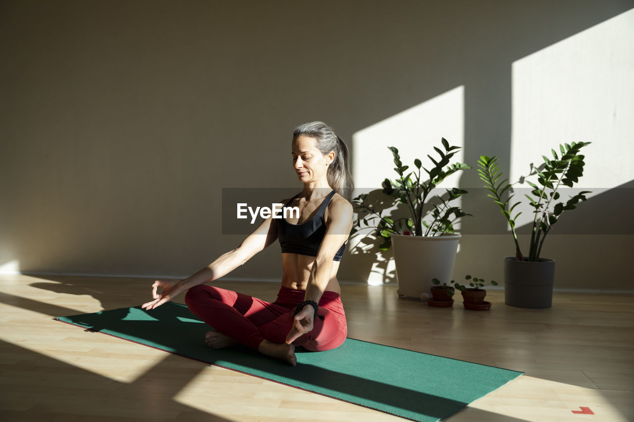 Yogini meditating on exercise mat in living room on sunny day
