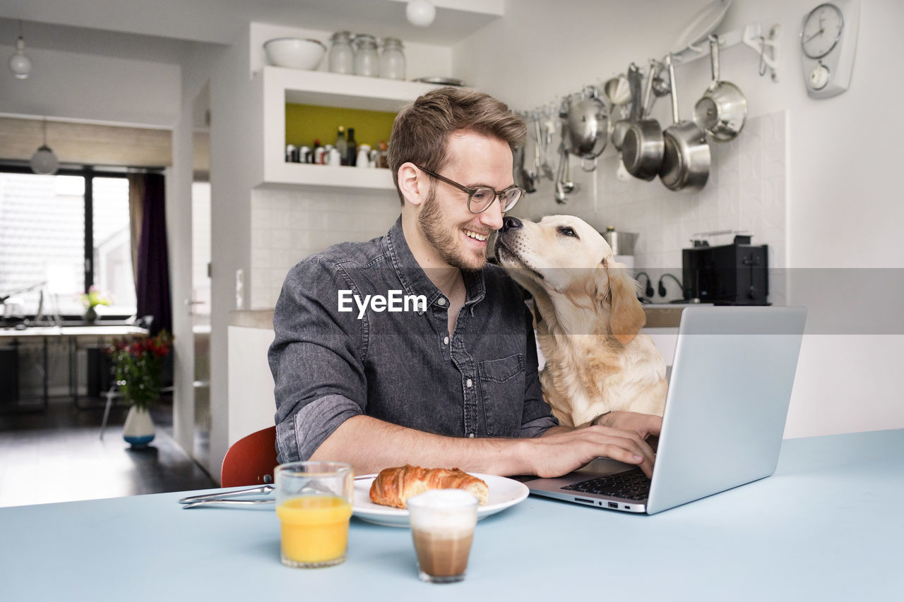 Smiling man with dog using laptop in kitchen at home