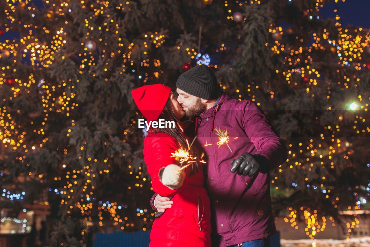 Couple kissing while holding sparkler against illuminated tree