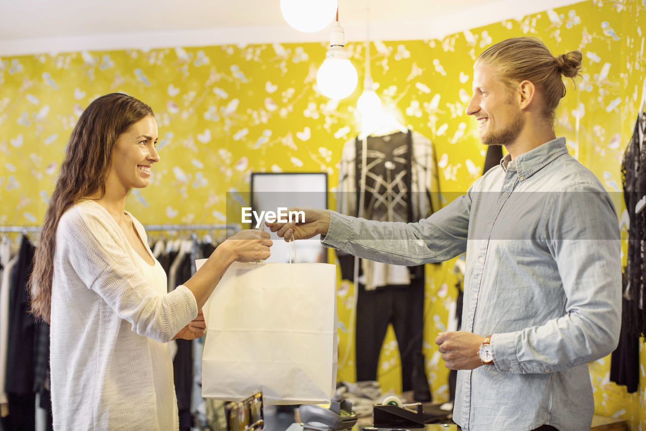 Happy female customer taking shopping bag from sales person at checkout in boutique