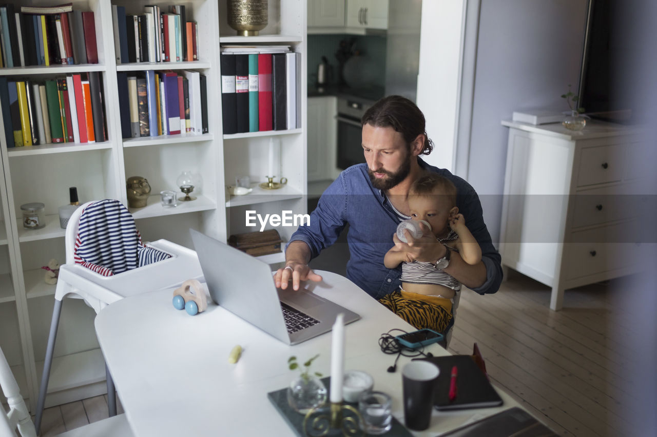 Mid adult father feeding baby boy while using laptop at home