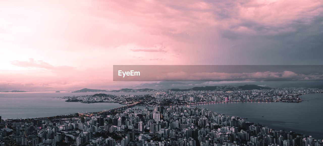 Aerial view of cityscape by sea against cloudy sky during sunset