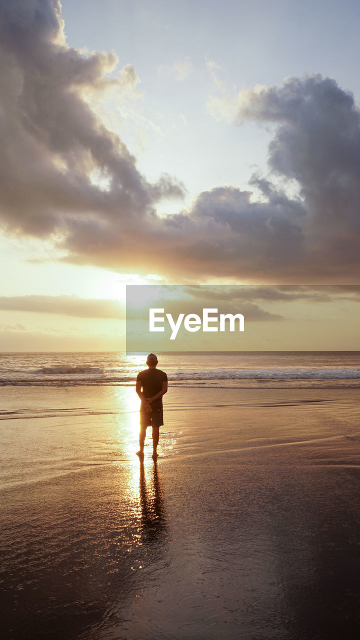 Rear view of man standing on beach against sky during sunset