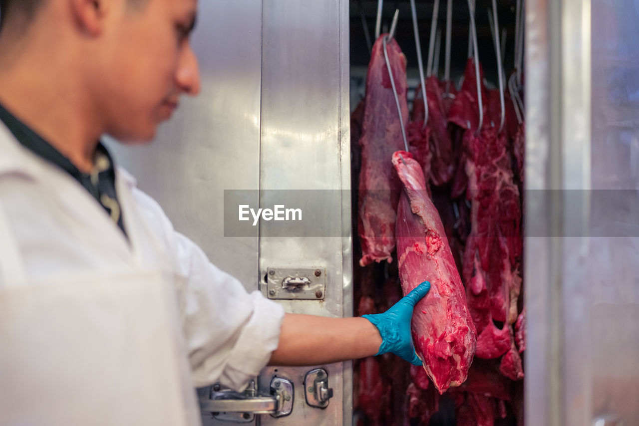 Man in latex gloves touching piece of raw meat hanging on hook behind metal door of refrigerator during work in butcher shop