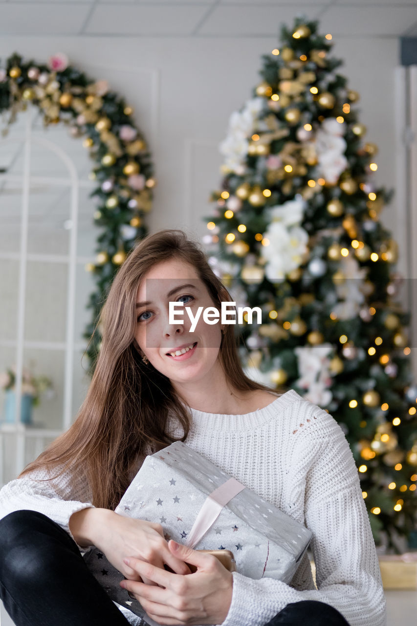 Girl in a white sweater sits with a christmas present on the background of a christmas tree. 