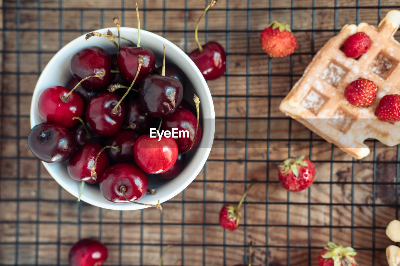 Fresh cherries in a bowl and strawberries with viennese waffles on a wooden table top view