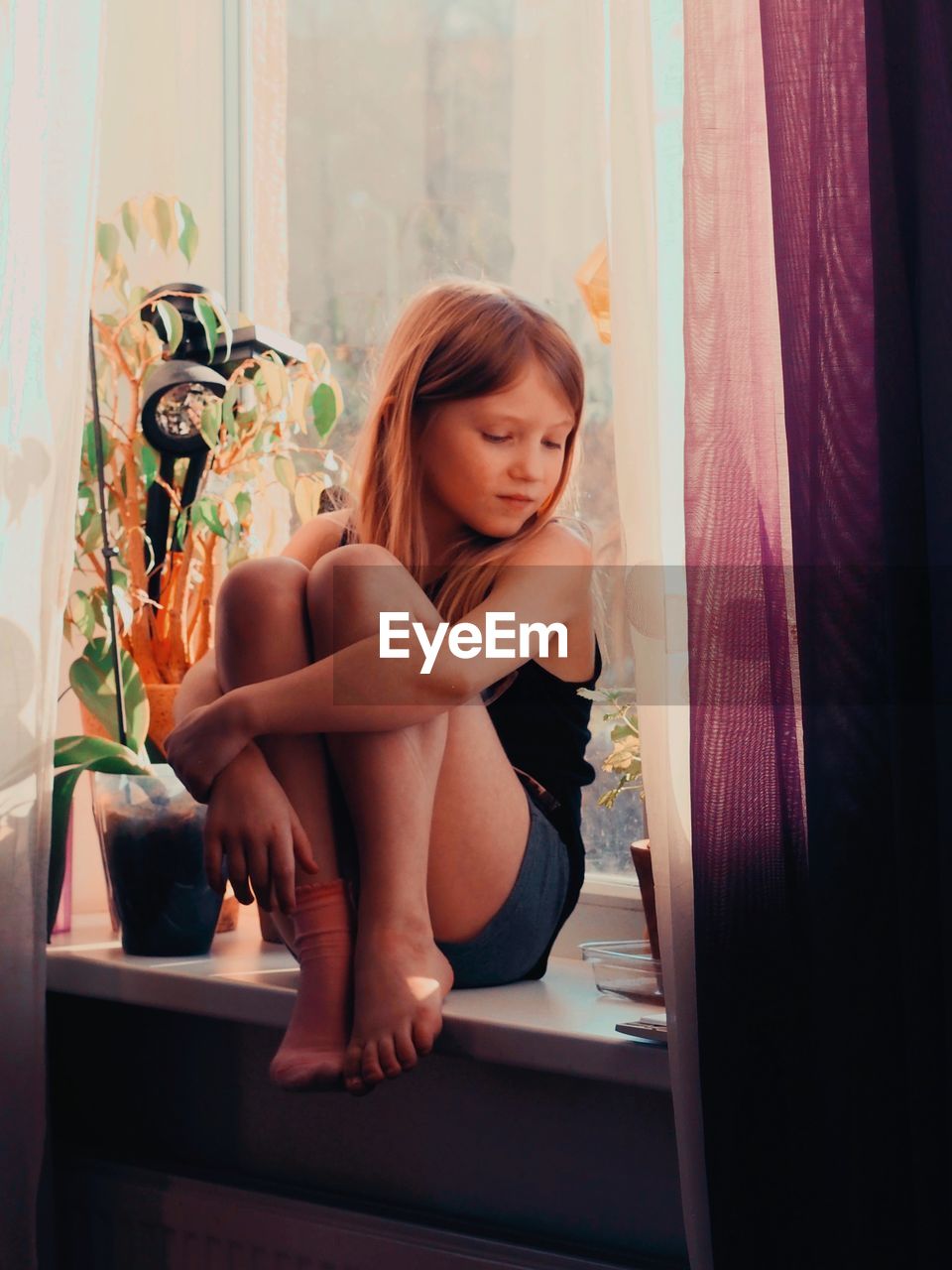 Young woman looking away while sitting on window at home