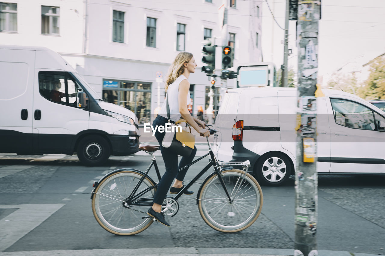 Full length of young businesswoman riding bicycle on road in city