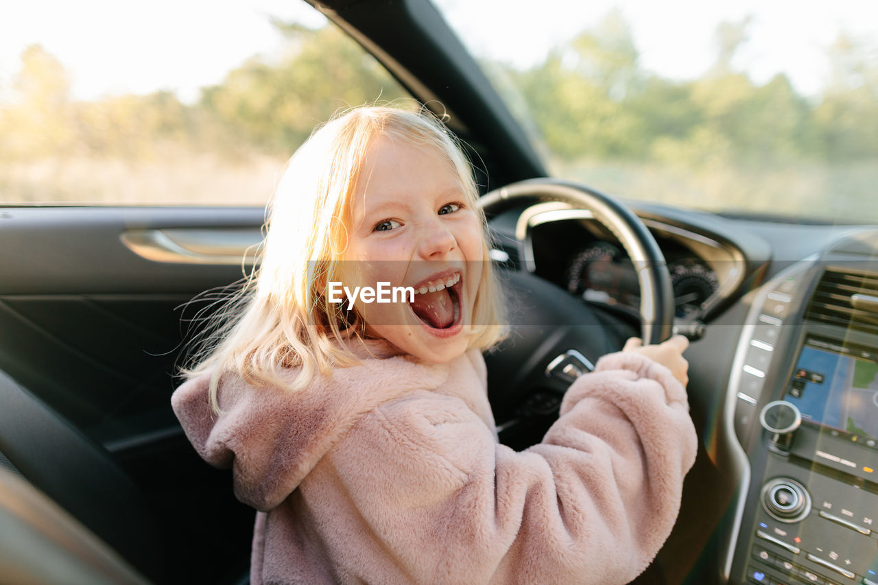 Side view of cheerful girl looking at camera sitting on driver seat of modern car with hands on steering wheel