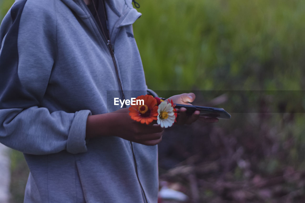 Midsection of woman holding flowers using smart phone while standing outdoors