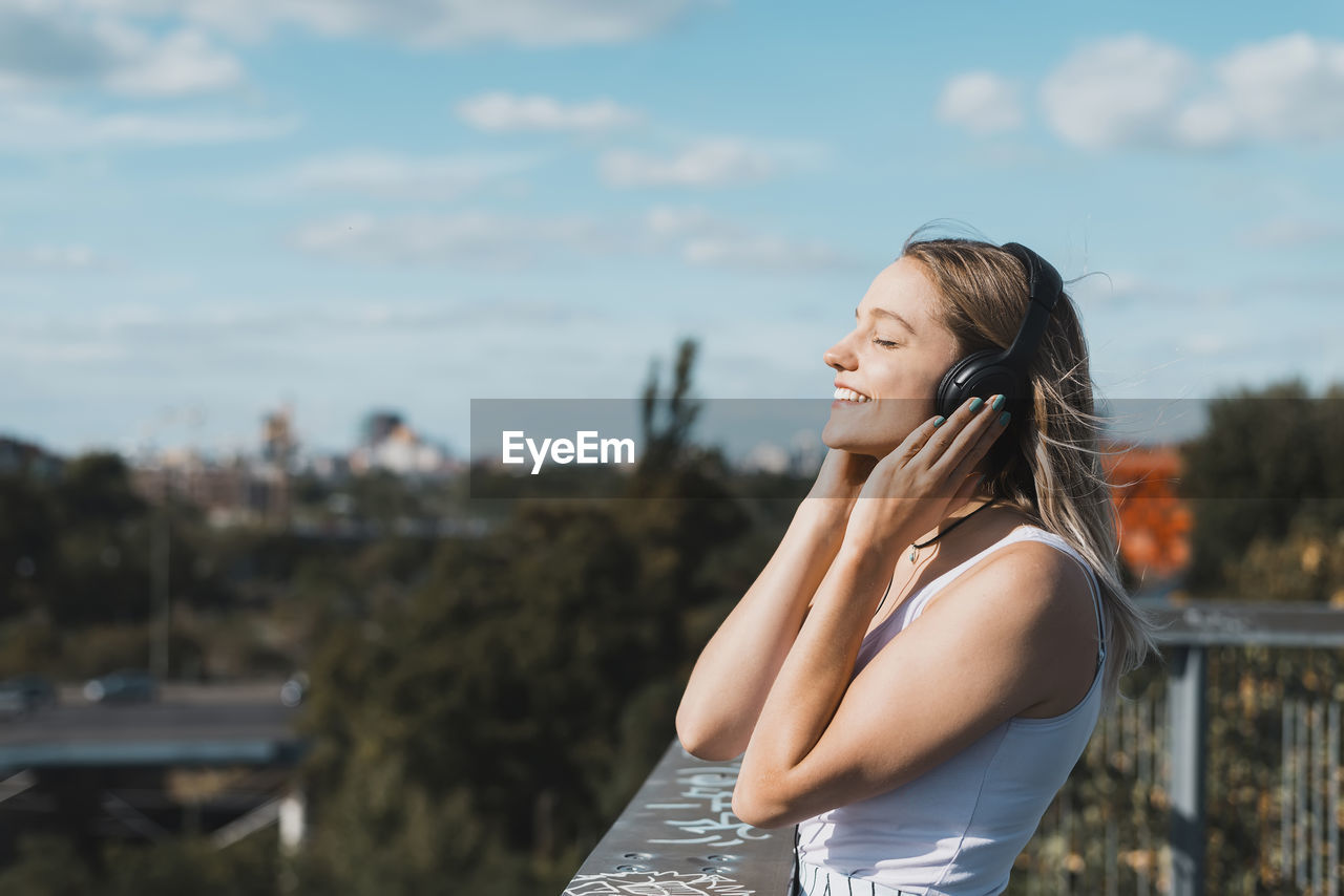 Side view of smiling young woman listening music by railing against sky