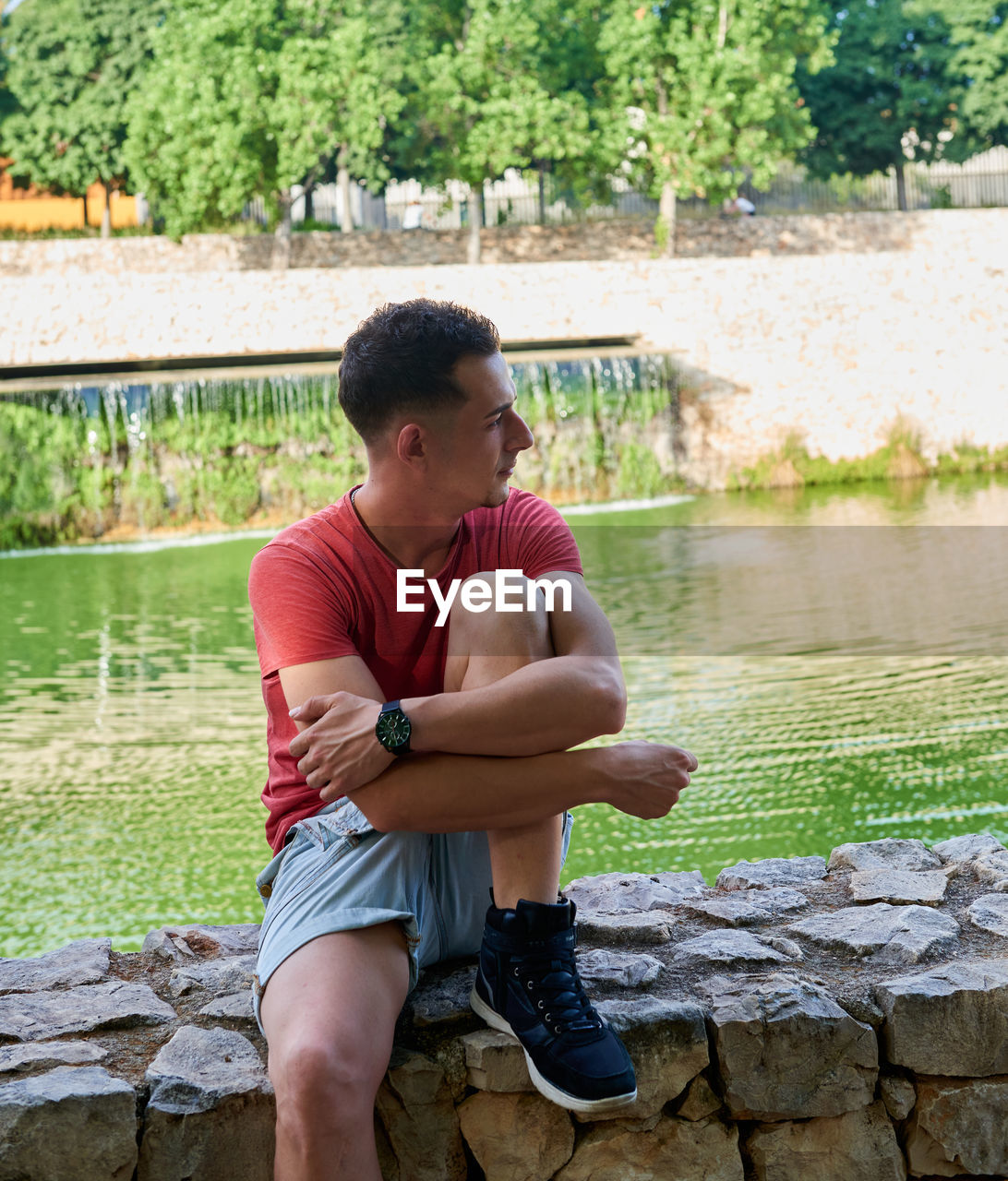 A young, attractive boy sitting on a stone wall next to a pond.