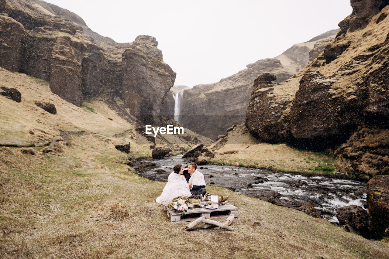 PEOPLE SITTING ON ROCKS AGAINST MOUNTAIN