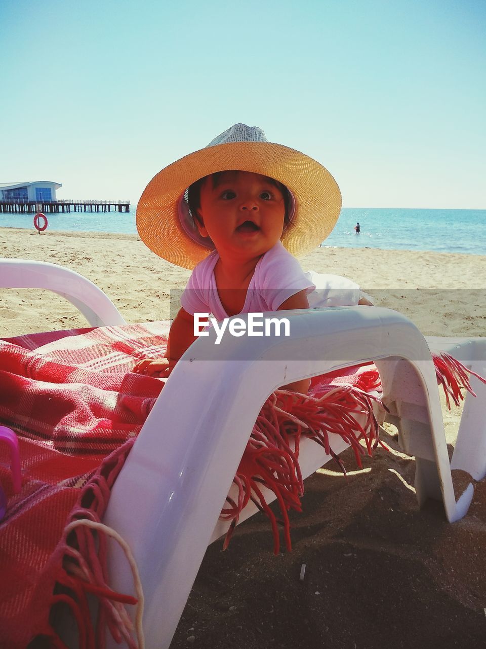 Cute baby girl wearing hat on chair at beach against sky