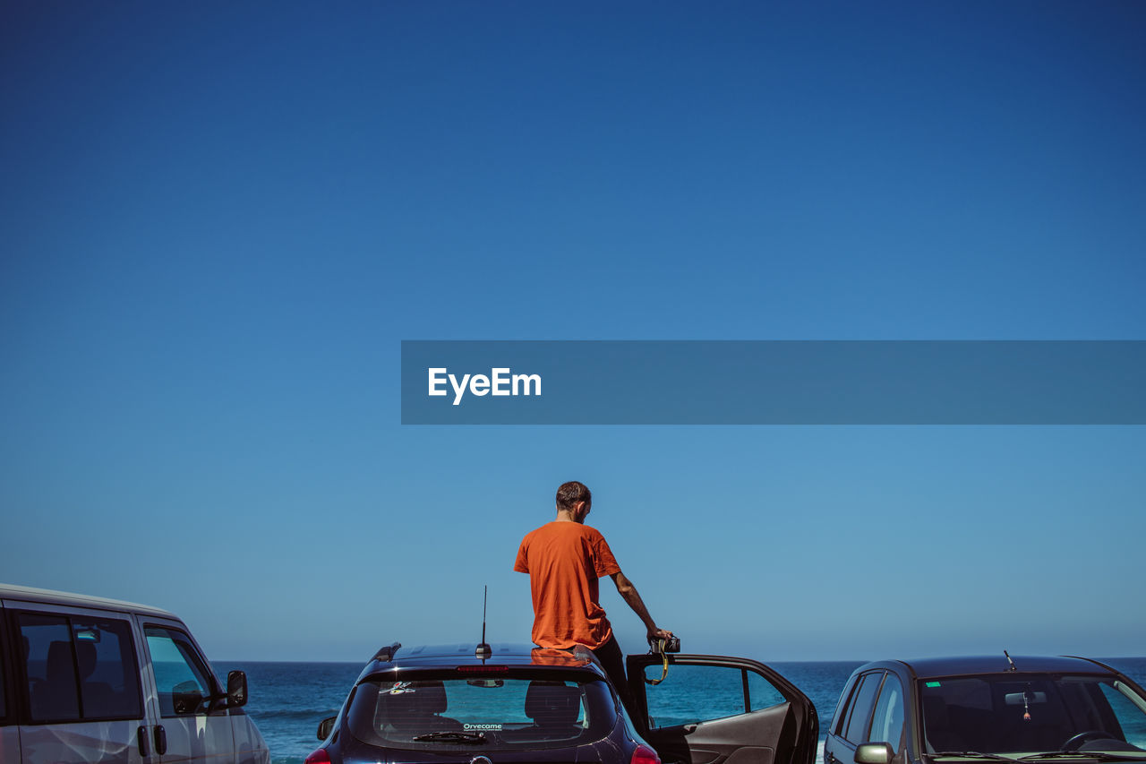 REAR VIEW OF MAN STANDING ON BOAT AGAINST CLEAR BLUE SKY