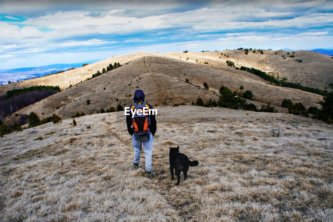 Young man from behind with backpack walking in hills with black dog against blue cloudy sky. hiking