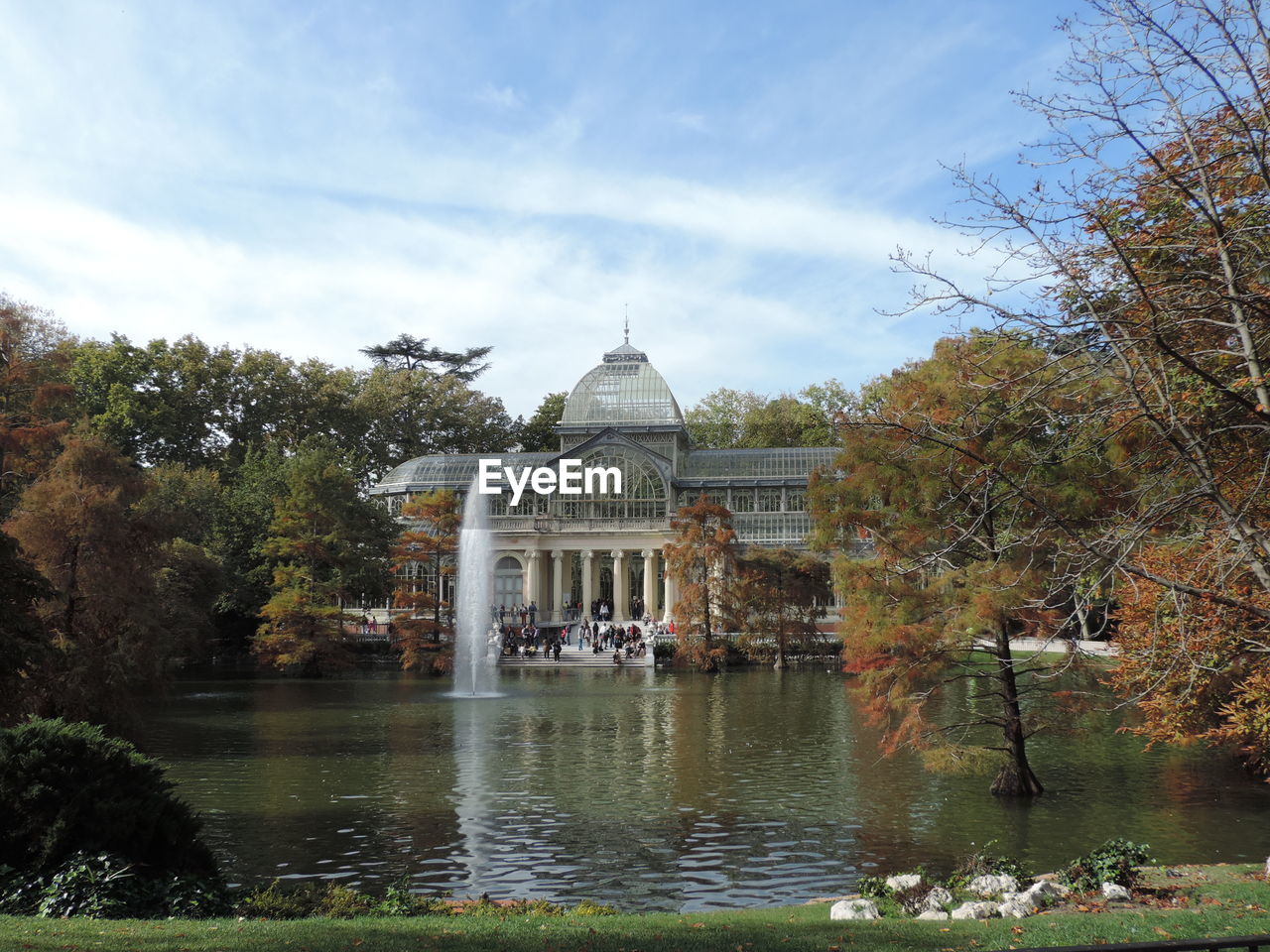 View of trees and fountain in river while tourists sightseeing famous place