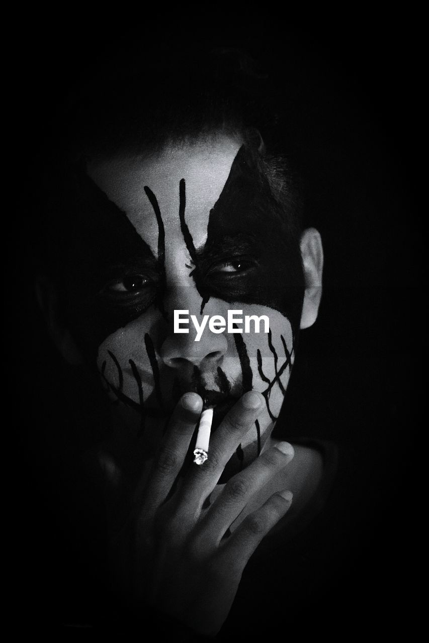 Close-up of man with zombie face paint smoking cigarette against black background