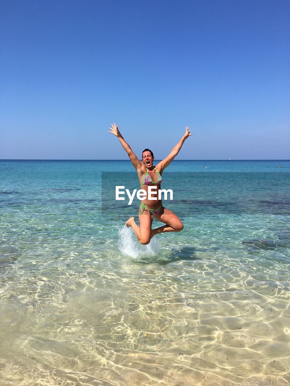 Cheerful young woman wearing bikini with arms raised jumping in sea against clear blue sky