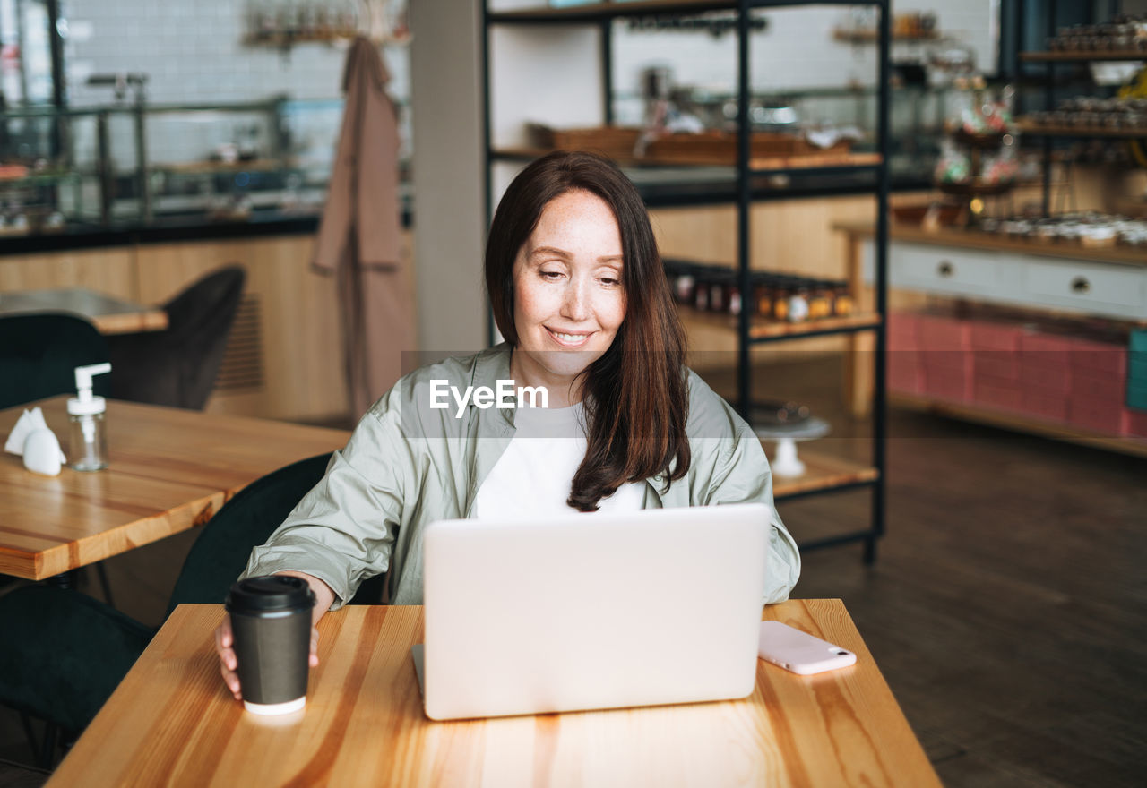 Adult smiling brunette business woman working on laptop using mobile phone in cafe