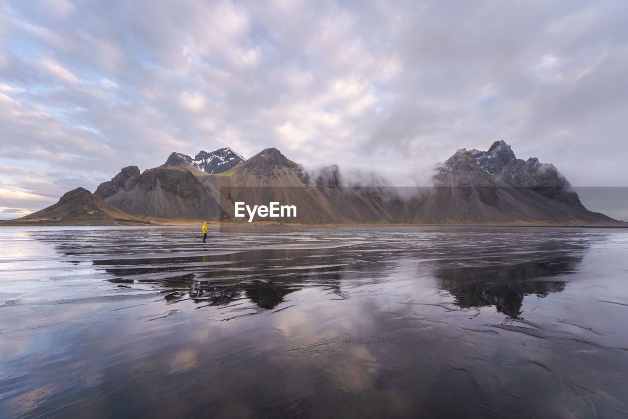 Anonymous tourist standing on wet shore near calm sea and rough mountains during majestic colorful sunset