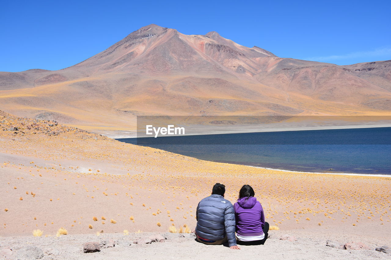 REAR VIEW OF FRIENDS SITTING ON DESERT AGAINST MOUNTAIN