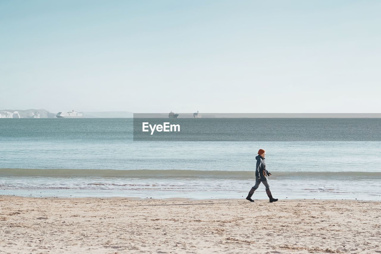 BOY STANDING ON BEACH AGAINST CLEAR SKY