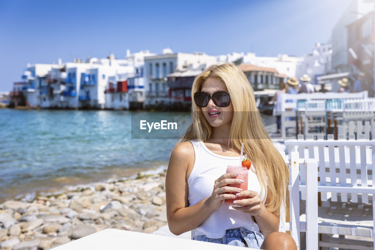 portrait of smiling young woman looking away while sitting at beach against sky