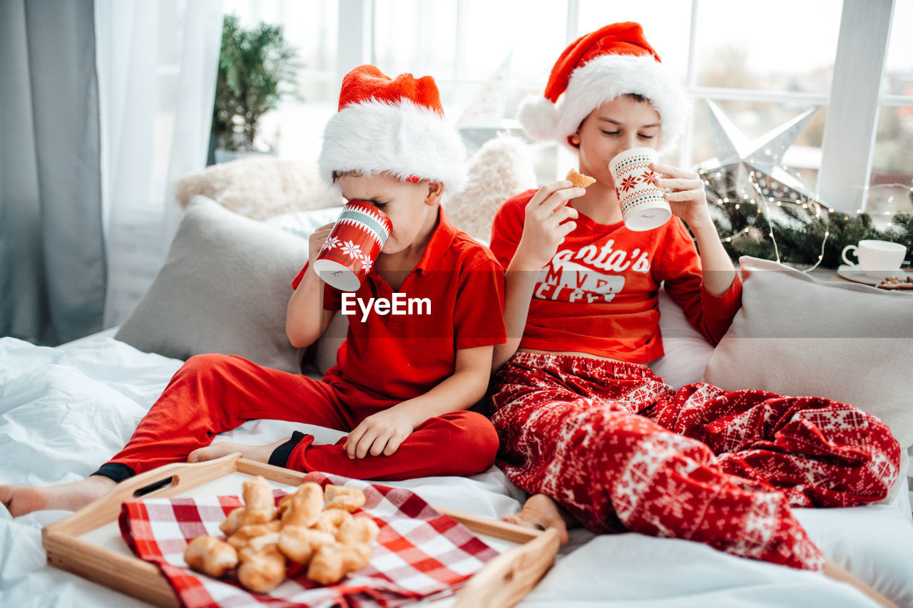 Cute kids drinking hot chocolate while sitting on bed at home