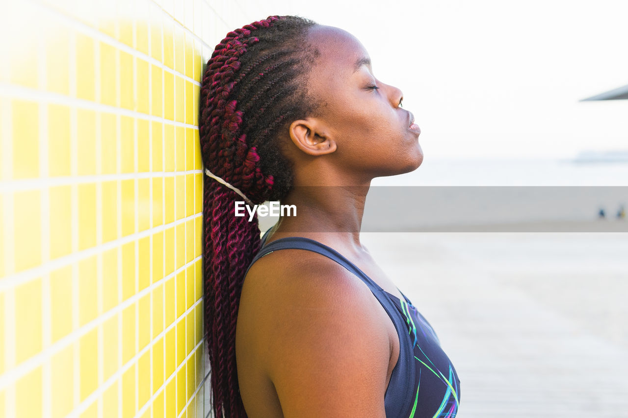 Fit black female athlete in sportswear resting leaning against yellow tiled wall after working out