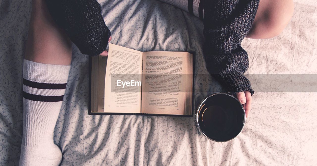 High angle view of woman reading with coffee cup on bed