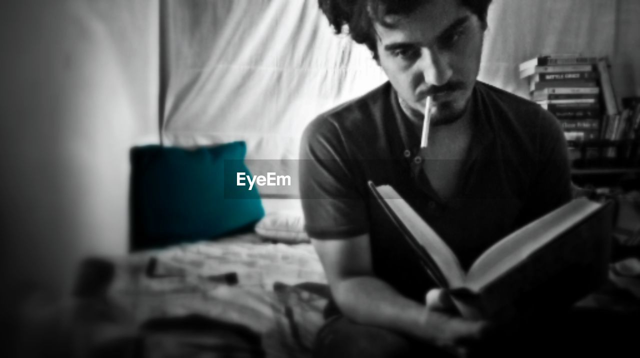 Young man reading book while smoking at home