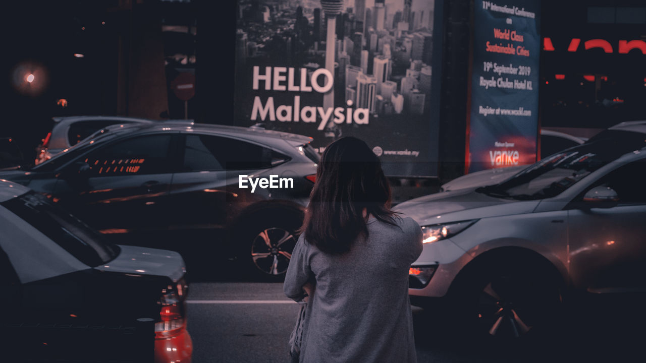 REAR VIEW OF WOMAN STANDING ON STREET IN CITY AT NIGHT