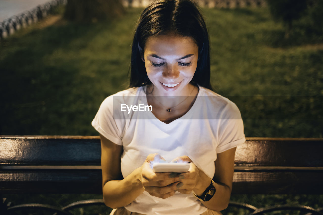 Happy woman using smart phone sitting on park bench at night