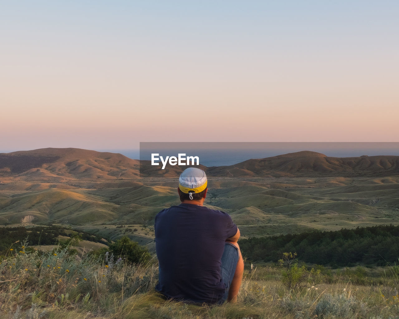 REAR VIEW OF MAN SITTING ON MOUNTAIN AGAINST CLEAR SKY