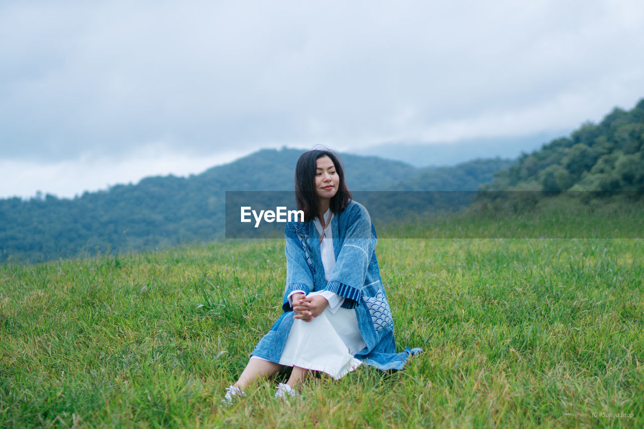 Woman sitting on grassy land against cloudy sky