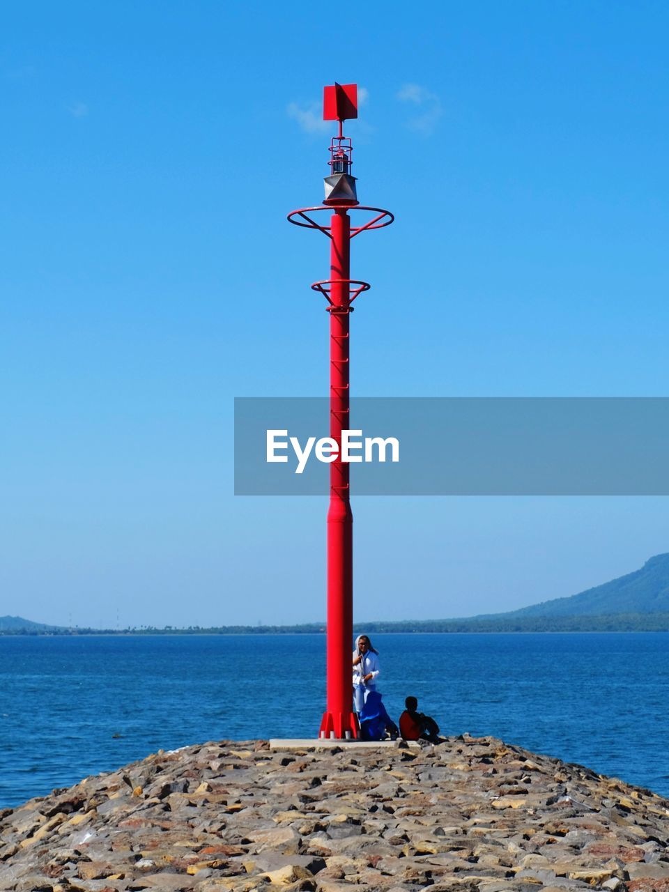 People relaxing by lighthouse on groyne by sea against clear blue sky