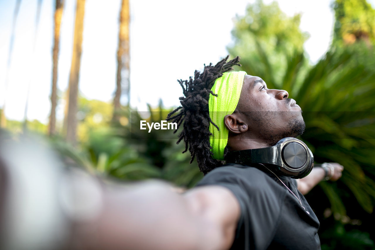 Young african american male athlete in sportswear with headphones on neck keeping open arms and looking away thoughtfully while standing in green tropical park