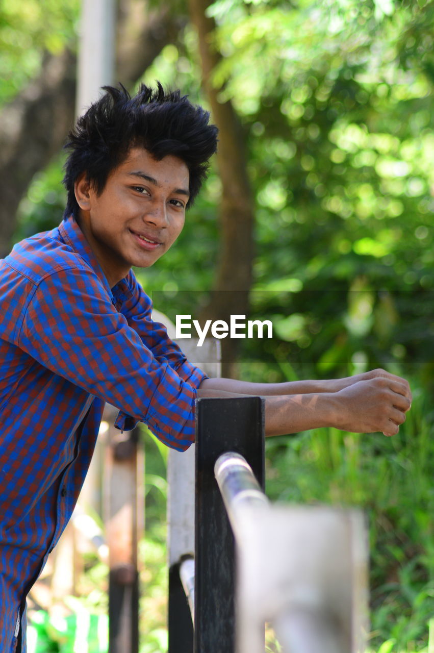 Portrait of young man leaning on railing against trees