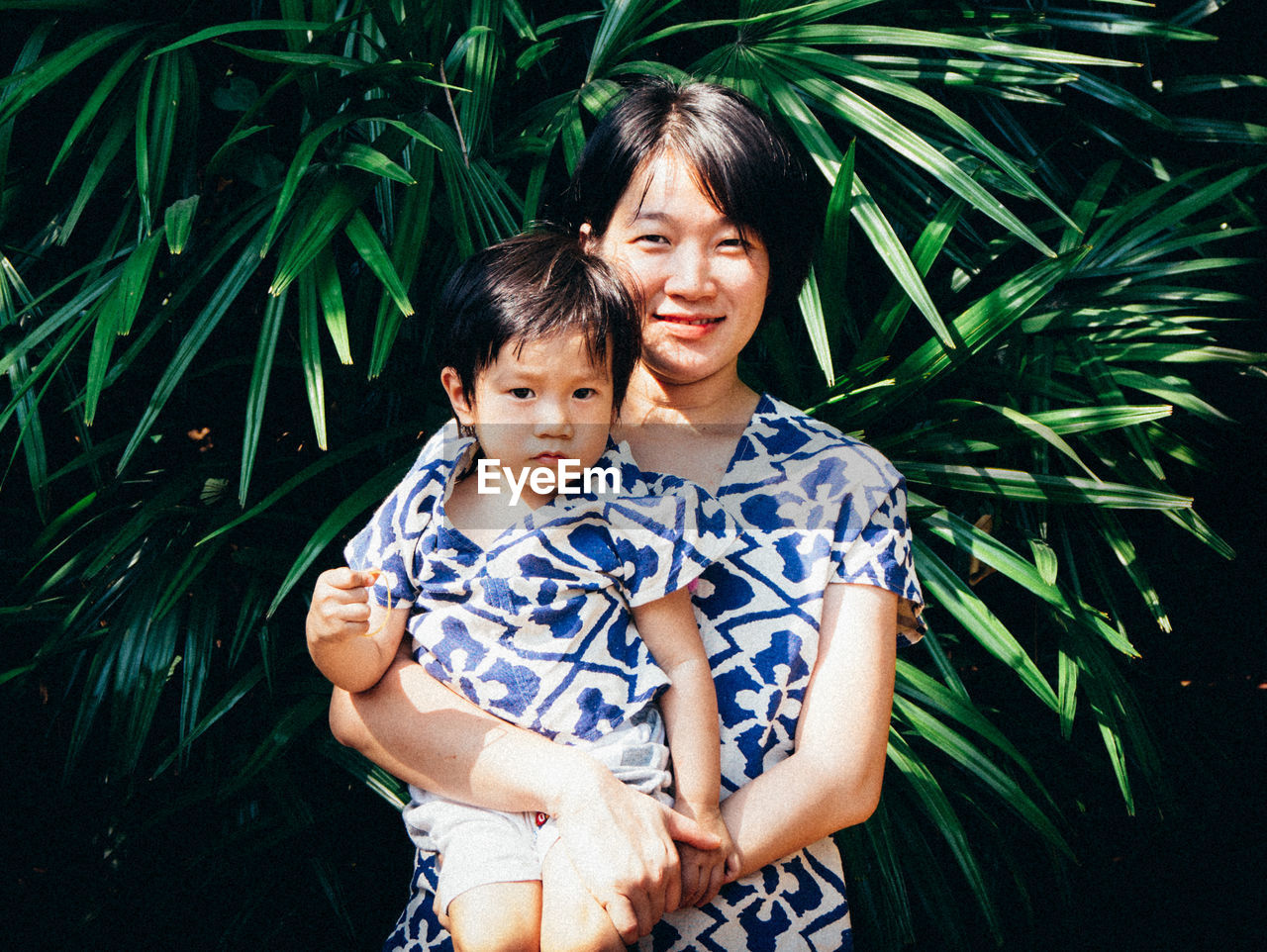 Portrait of smiling mother carrying cute son while standing against plants