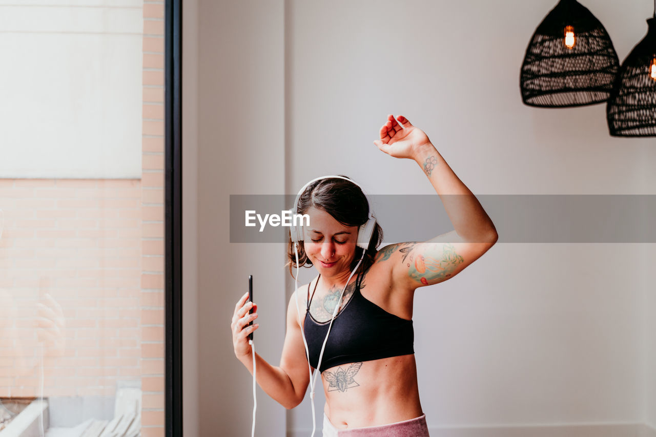 Happy muscular woman listening to music on mobile phone and headset in the gym. dancing by window