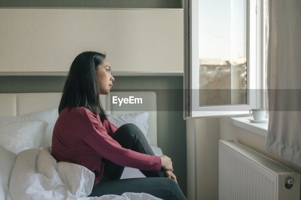 SIDE VIEW OF WOMAN SITTING ON BED AT HOME